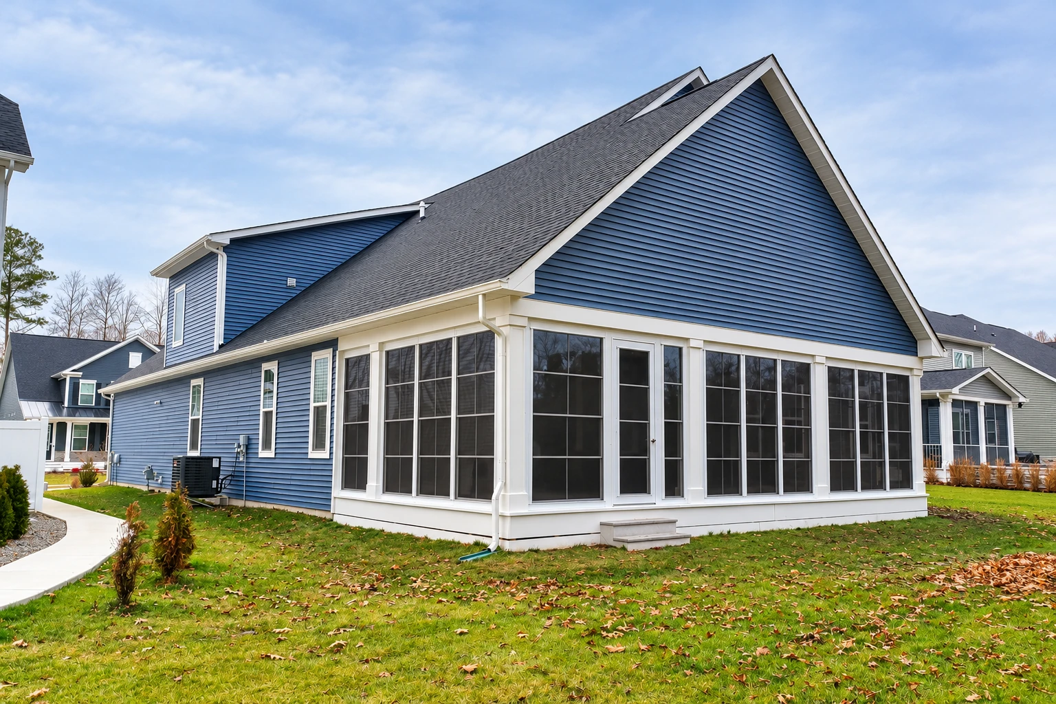 A blue-sided house has a large enclosed porch with white framing and dark screened panels overlooking a green lawn.