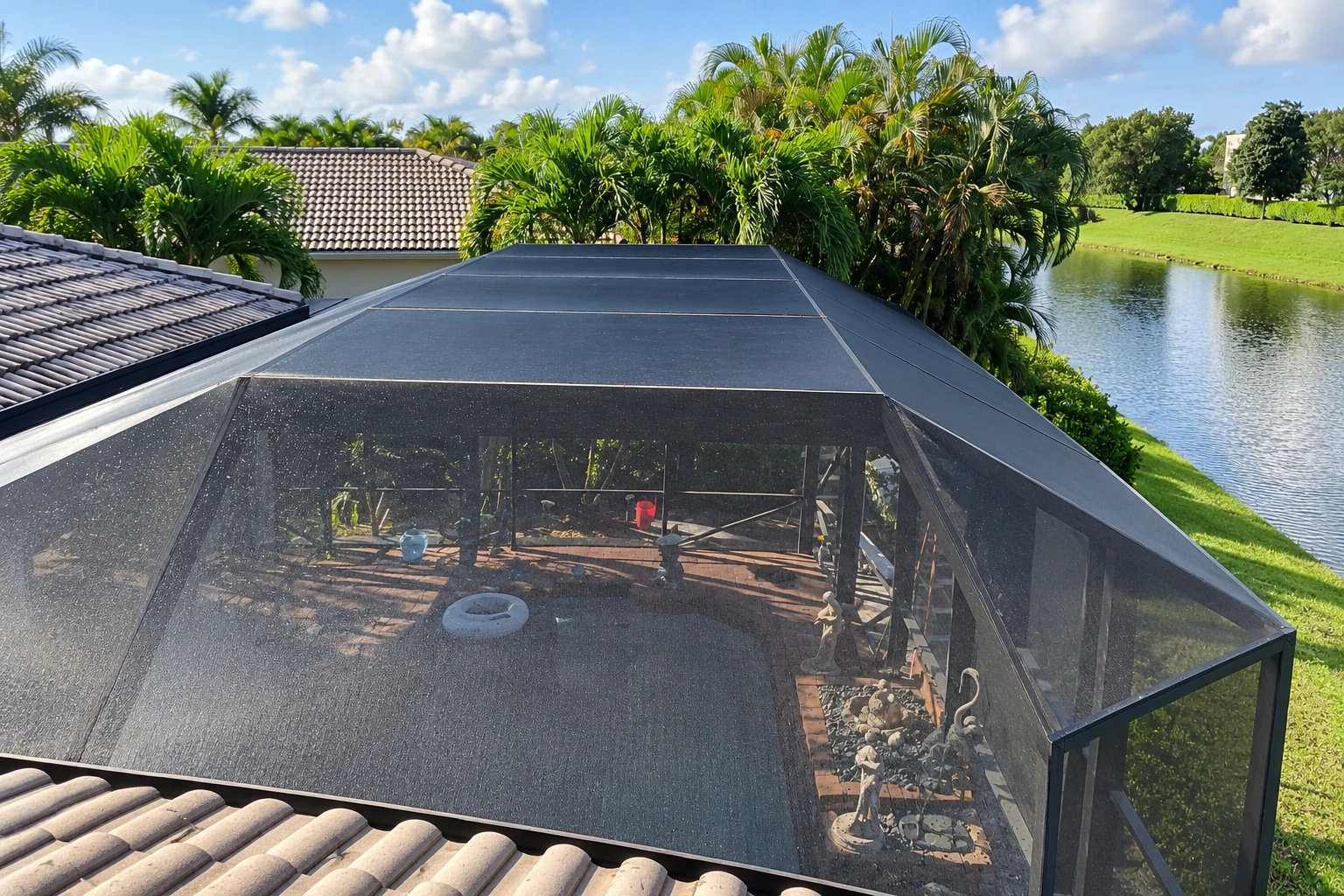 Black mesh screen enclosure over a patio beside a tiled-roof house, palm trees and a pond.