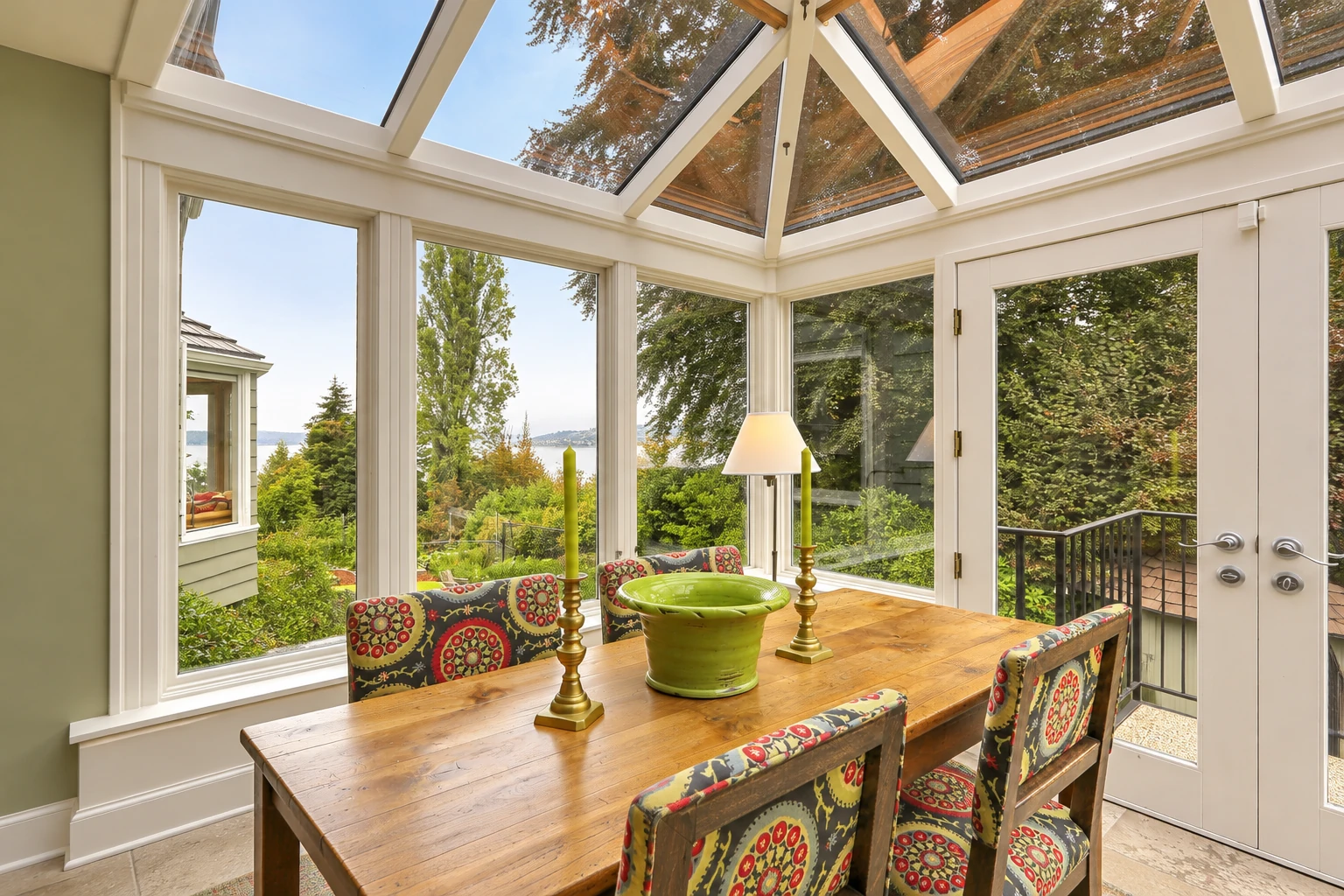 A solarium with white-framed glass walls and a peaked glass roof, furnished with a long wooden dining table and patterned chairs.