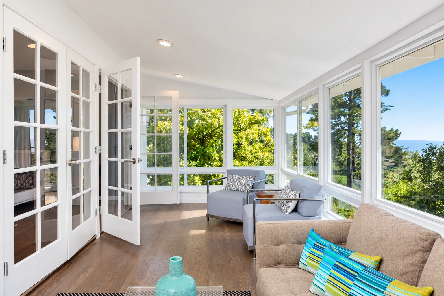 A bright sunroom with white-framed windows and open French doors, furnished with sofas and overlooking leafy greenery.