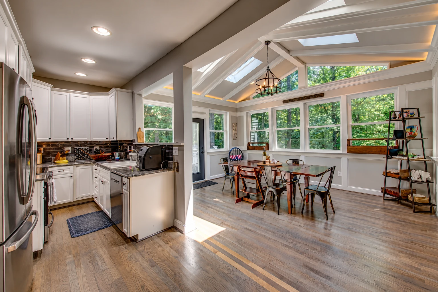 A sunroom dining area with white-trimmed windows and skylights connects to a kitchen with white cabinets and stainless steel appliances.