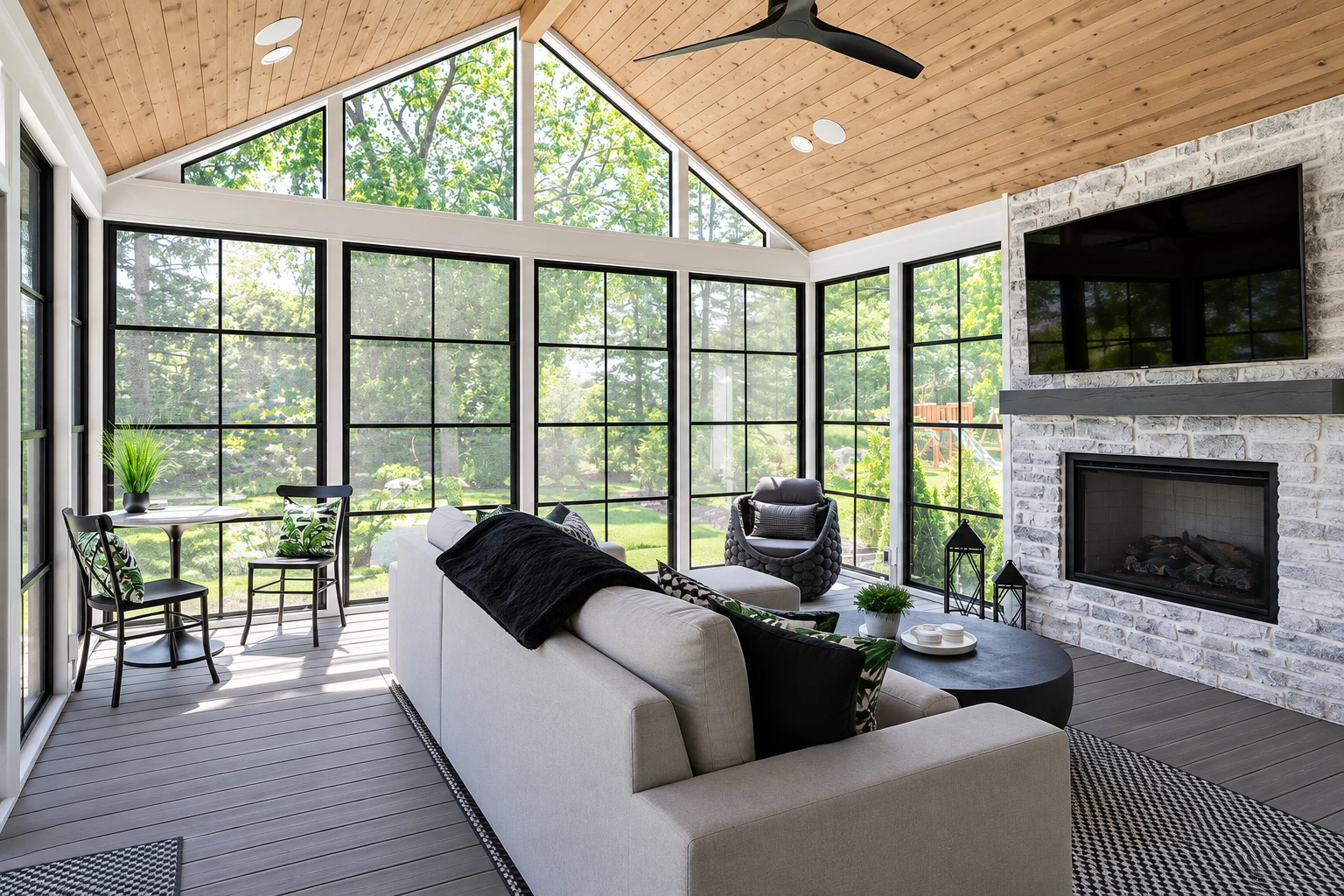 A furnished sunroom has tall black-framed windows, a vaulted wood ceiling, and a stone fireplace.