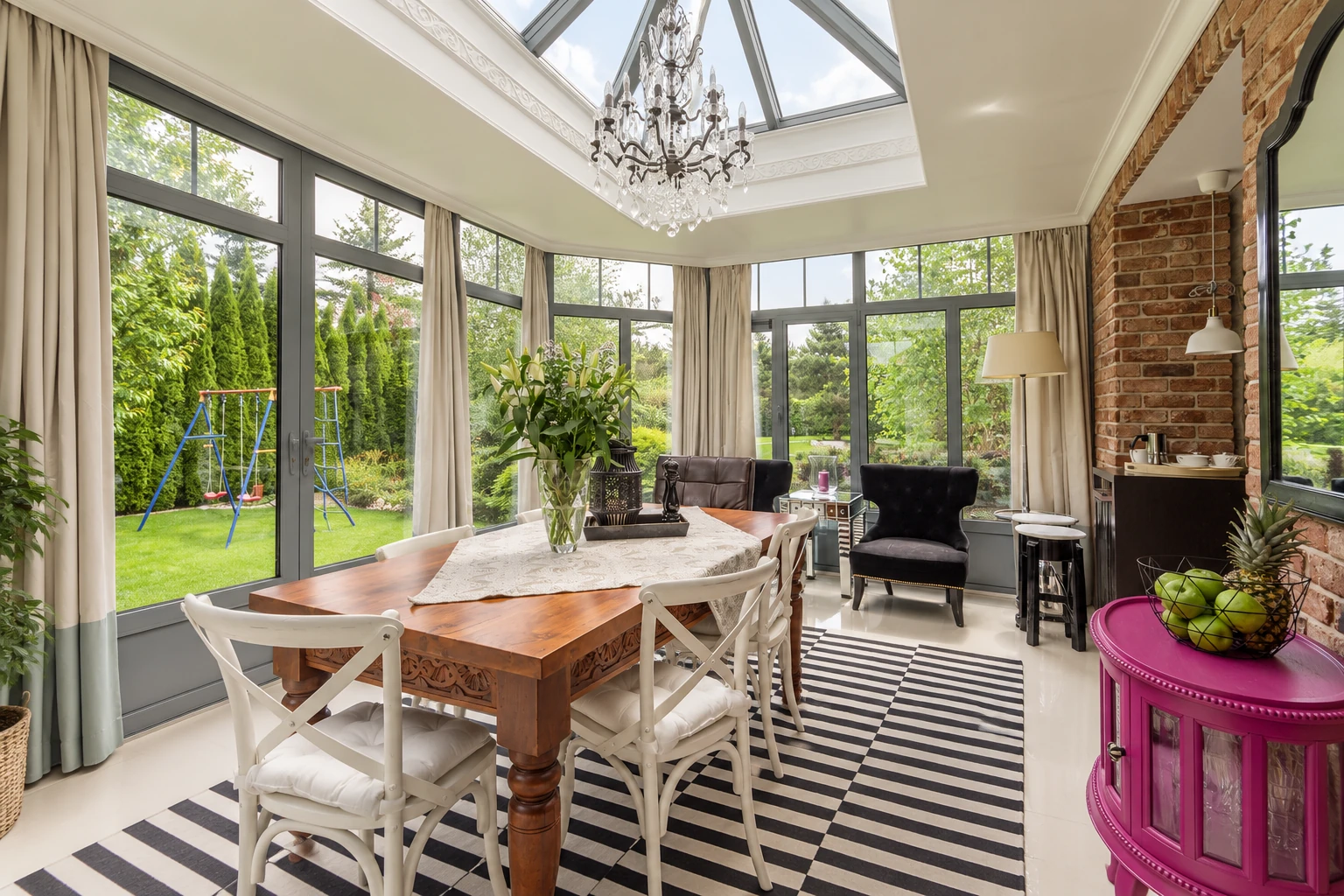 A bright three-season sunroom has tall windows, a glass skylight, a dining table, and views of a garden.